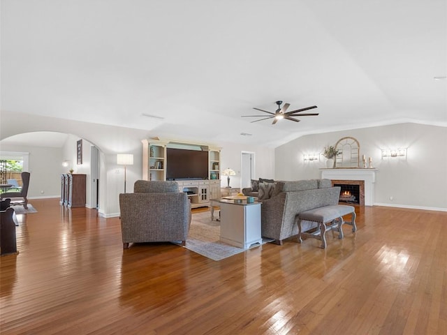 living area with ceiling fan, arched walkways, vaulted ceiling, a fireplace with flush hearth, and wood finished floors