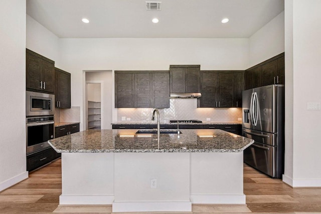 181 Benedum Way, Liberty Hill, TX 78642 - kitchen featuring appliances with stainless steel finishes, dark stone counters, backsplash, a kitchen island with sink, and dark brown cabinetry