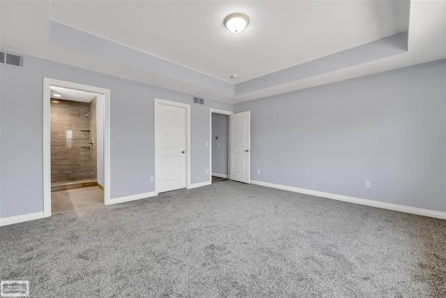 unfurnished bedroom with a tray ceiling, ensuite bathroom, and dark colored carpet