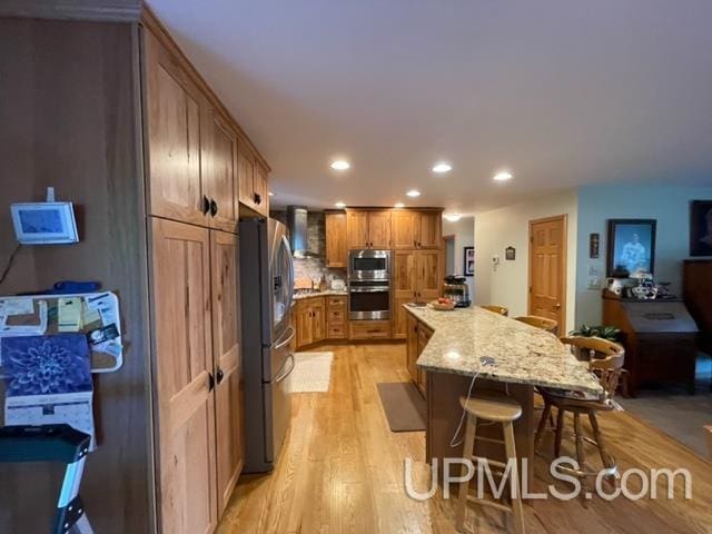 kitchen featuring wall chimney range hood, a center island, a breakfast bar, appliances with stainless steel finishes, and light hardwood / wood-style flooring