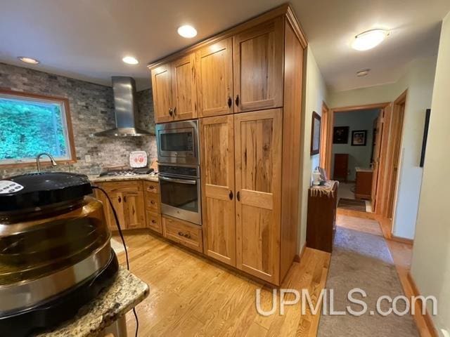 kitchen with backsplash, appliances with stainless steel finishes, light wood-type flooring, and wall chimney range hood