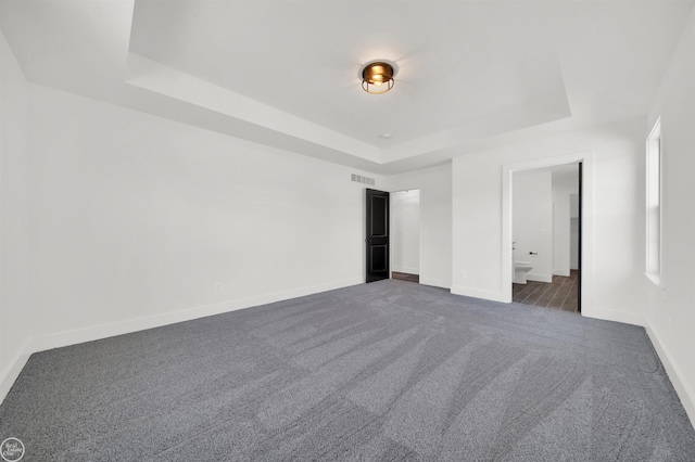 unfurnished bedroom with a raised ceiling, ensuite bathroom, and dark carpet