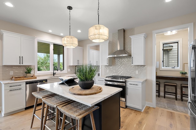 kitchen with a center island, wall chimney range hood, light wood-type flooring, appliances with stainless steel finishes, and white cabinetry