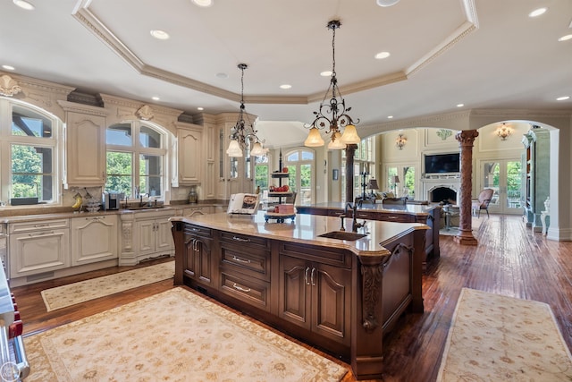 kitchen featuring pendant lighting, a kitchen island with sink, dark hardwood / wood-style floors, a tray ceiling, and ornate columns