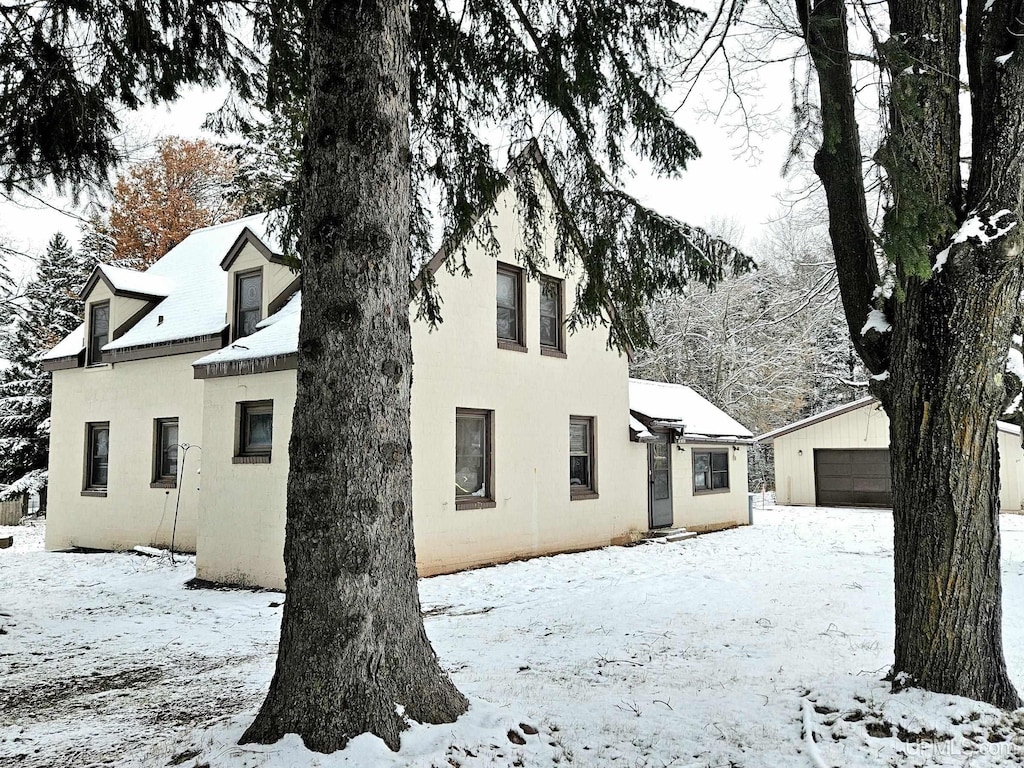 snow covered house with stucco siding, a detached garage, and an outbuilding