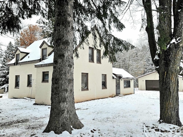 snow covered house with stucco siding, a detached garage, and an outbuilding