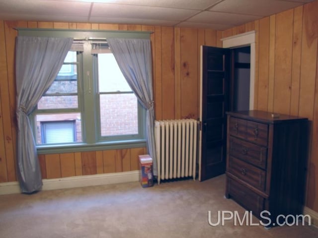 carpeted bedroom featuring wooden walls, multiple windows, a drop ceiling, and radiator