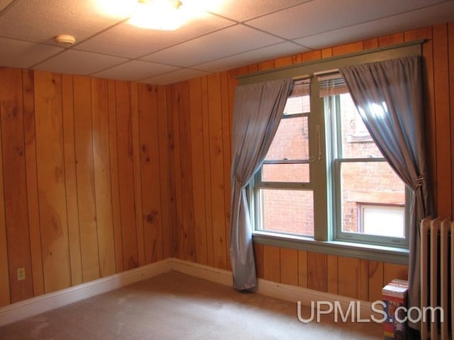 carpeted empty room with baseboards, a drop ceiling, wooden walls, and radiator heating unit