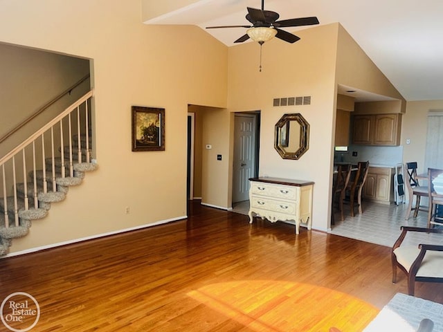 unfurnished living room with wood finished floors, visible vents, a ceiling fan, high vaulted ceiling, and stairs