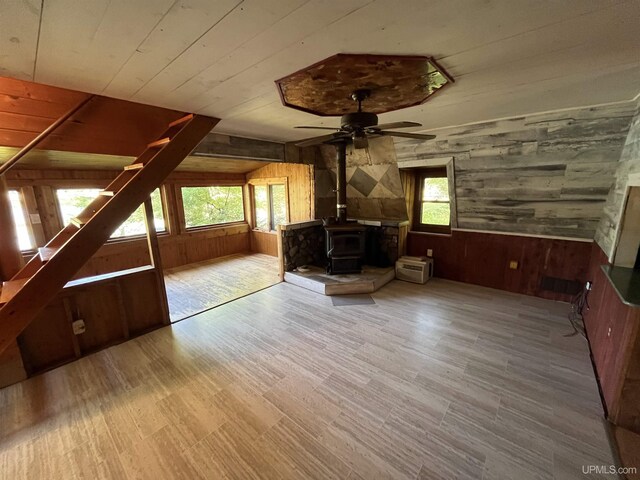 unfurnished living room with ceiling fan, wooden walls, a wood stove, and a healthy amount of sunlight