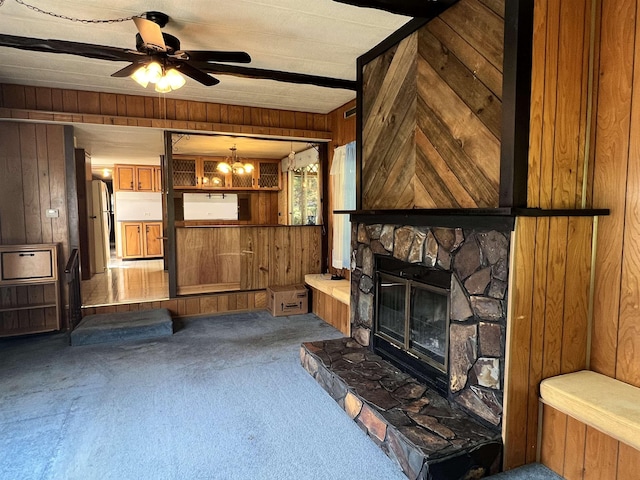 unfurnished living room featuring ceiling fan with notable chandelier, carpet floors, a stone fireplace, and wood walls