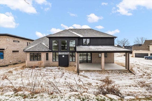 snow covered rear of property with a patio, brick siding, and roof with shingles