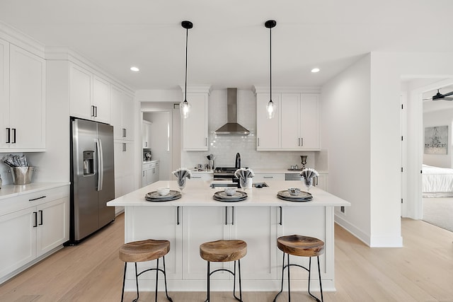 kitchen featuring a center island with sink, wall chimney exhaust hood, decorative light fixtures, stainless steel fridge with ice dispenser, and light wood-type flooring