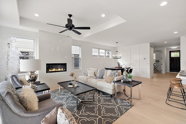 living room with ceiling fan with notable chandelier, a raised ceiling, and light hardwood / wood-style flooring