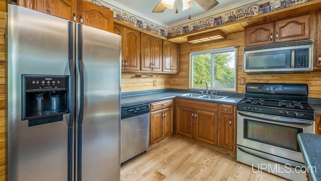 kitchen with light hardwood / wood-style floors, sink, wood walls, stainless steel appliances, and ceiling fan