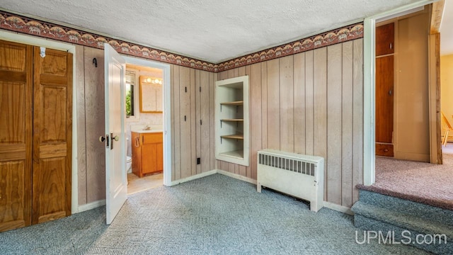 unfurnished bedroom with connected bathroom, wood walls, radiator heating unit, a textured ceiling, and carpet