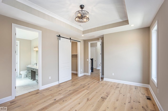 unfurnished bedroom featuring light hardwood / wood-style flooring, a tray ceiling, a notable chandelier, ensuite bath, and a barn door