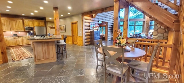 kitchen with a kitchen island with sink, log walls, light brown cabinetry, light stone countertops, and stainless steel microwave