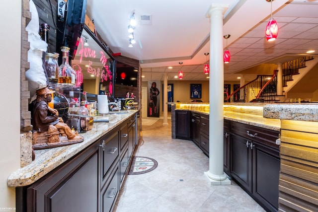 bar with light stone counters, a paneled ceiling, decorative light fixtures, dark brown cabinetry, and ornate columns