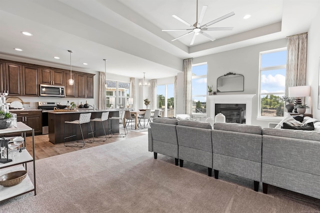 living room with ceiling fan with notable chandelier, light wood-type flooring, plenty of natural light, and a tray ceiling