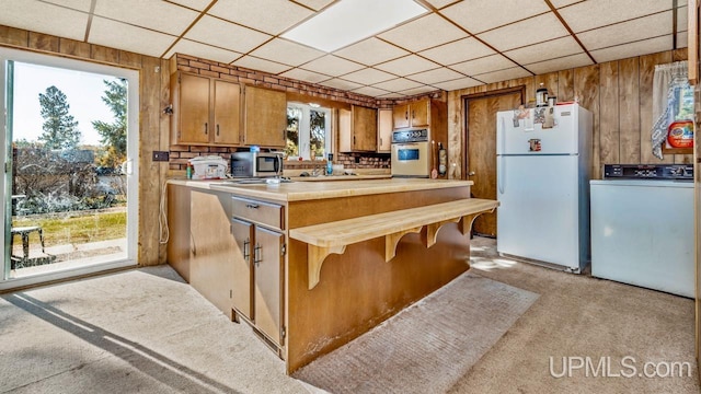 kitchen with brown cabinets, wood walls, light carpet, white appliances, and washer / clothes dryer