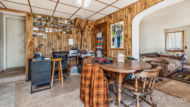 carpeted dining area featuring arched walkways, wooden walls, and a drop ceiling