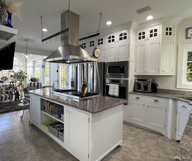kitchen featuring stainless steel appliances, island range hood, dark stone counters, a kitchen island with sink, and white cabinetry