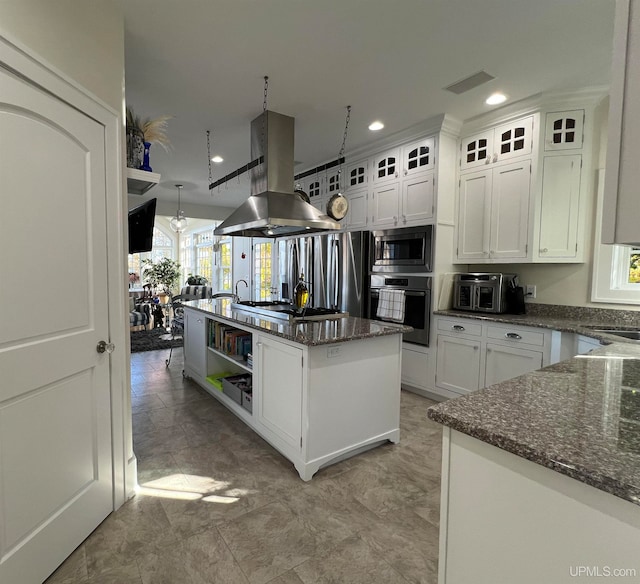 kitchen with appliances with stainless steel finishes, island range hood, white cabinetry, dark stone countertops, and a center island with sink