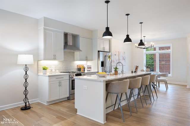 kitchen featuring wall chimney range hood, appliances with stainless steel finishes, white cabinetry, light hardwood / wood-style floors, and a center island with sink