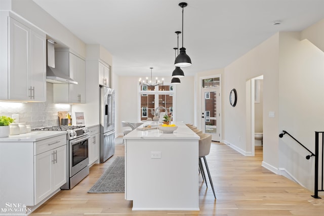 kitchen with wall chimney range hood, hanging light fixtures, white cabinetry, a kitchen island with sink, and premium appliances