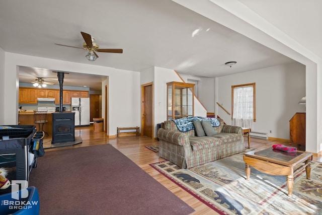 living room featuring light hardwood / wood-style floors, a wood stove, a baseboard heating unit, and ceiling fan