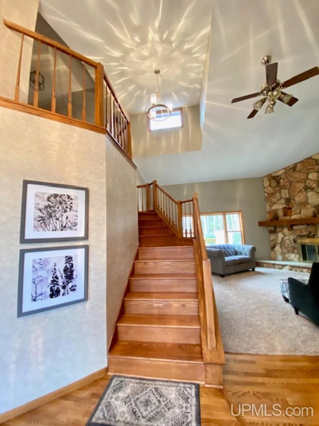 stairway with a stone fireplace, ceiling fan, high vaulted ceiling, and wood-type flooring