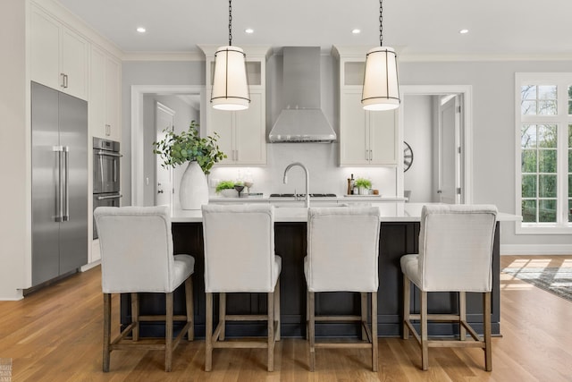 kitchen featuring appliances with stainless steel finishes, wall chimney exhaust hood, light countertops, decorative backsplash, and light wood-style floors