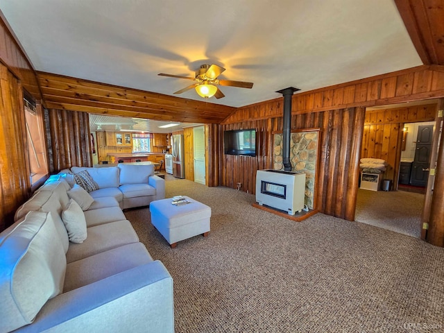 carpeted living area featuring wooden walls, ceiling fan, a wood stove, vaulted ceiling, and a wealth of natural light