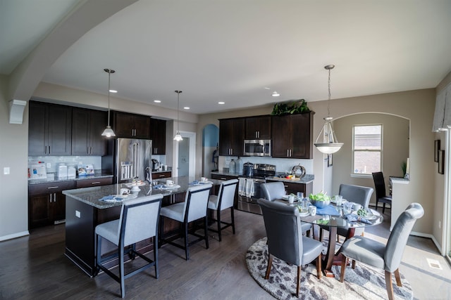 kitchen featuring dark hardwood / wood-style flooring, a kitchen island with sink, dark brown cabinets, appliances with stainless steel finishes, and decorative light fixtures