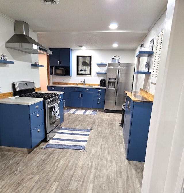 kitchen with wall chimney range hood, sink, light wood-type flooring, blue cabinetry, and appliances with stainless steel finishes