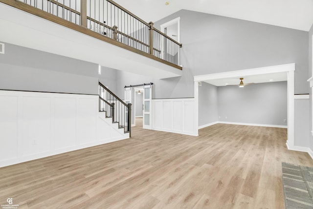 unfurnished living room with ceiling fan, a barn door, high vaulted ceiling, and light hardwood / wood-style flooring