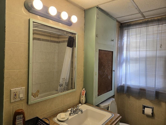 bathroom with a paneled ceiling, toilet, walk in shower, tile walls, and sink