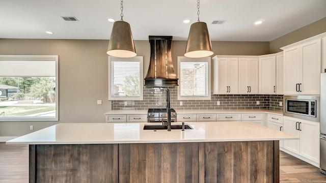 kitchen with hanging light fixtures, premium range hood, white cabinets, and a kitchen island with sink