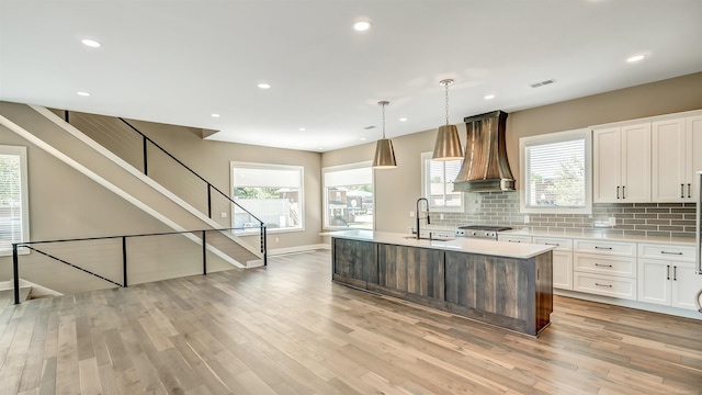 kitchen with light wood finished floors, custom range hood, a kitchen island with sink, light countertops, and a sink