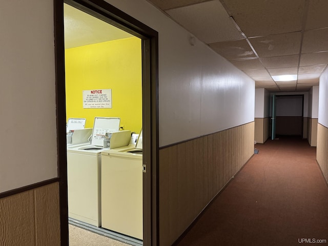 hallway with a paneled ceiling, washer and dryer, wooden walls, and light colored carpet