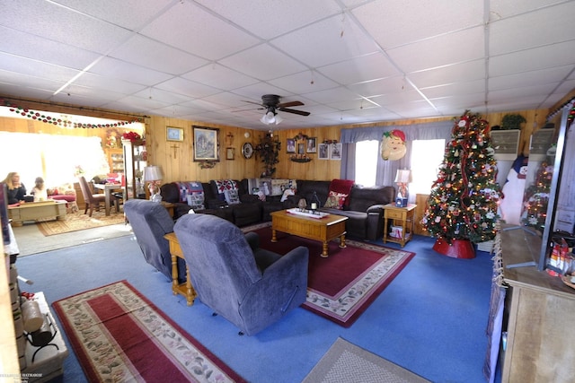 carpeted living room featuring a paneled ceiling, wooden walls, and ceiling fan