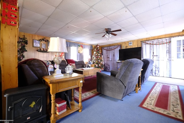 carpeted living room featuring a drop ceiling, wooden walls, and ceiling fan