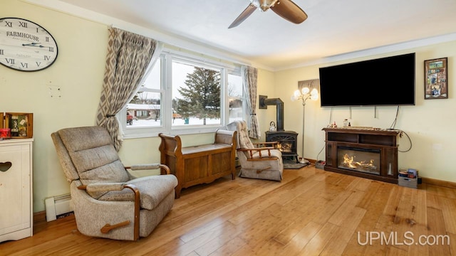 living area featuring a wood stove, light hardwood / wood-style flooring, ceiling fan, ornamental molding, and a baseboard radiator