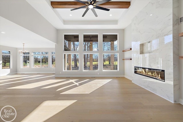 unfurnished living room with ceiling fan with notable chandelier, a tray ceiling, light hardwood / wood-style flooring, a premium fireplace, and a high ceiling