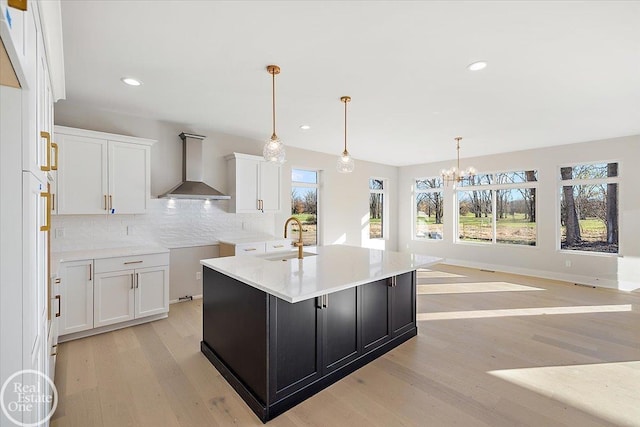 kitchen featuring white cabinetry, sink, wall chimney range hood, a center island with sink, and light wood-type flooring