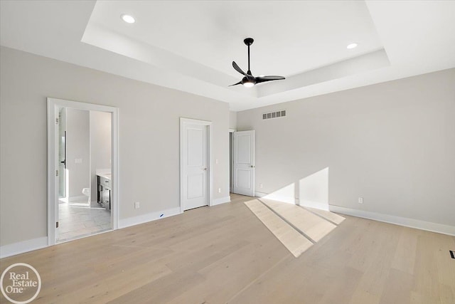 unfurnished bedroom with ensuite bathroom, light hardwood / wood-style flooring, ceiling fan, and a tray ceiling