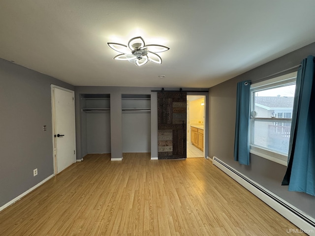 unfurnished bedroom featuring a baseboard heating unit, ensuite bathroom, a barn door, light hardwood / wood-style floors, and multiple closets