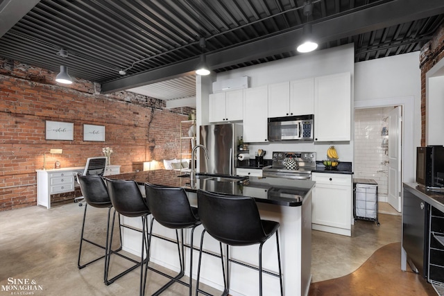 kitchen featuring white cabinetry, brick wall, a breakfast bar area, a kitchen island with sink, and appliances with stainless steel finishes