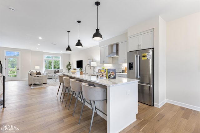 kitchen with a kitchen bar, high end refrigerator, wall chimney exhaust hood, white cabinetry, and an island with sink